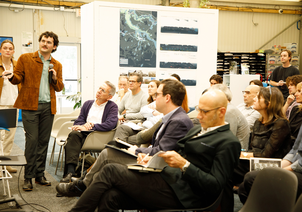 Two students stand at the left presenting a project that is off view to a group of faculty, experts, and fellow students seated in rows of chairs.